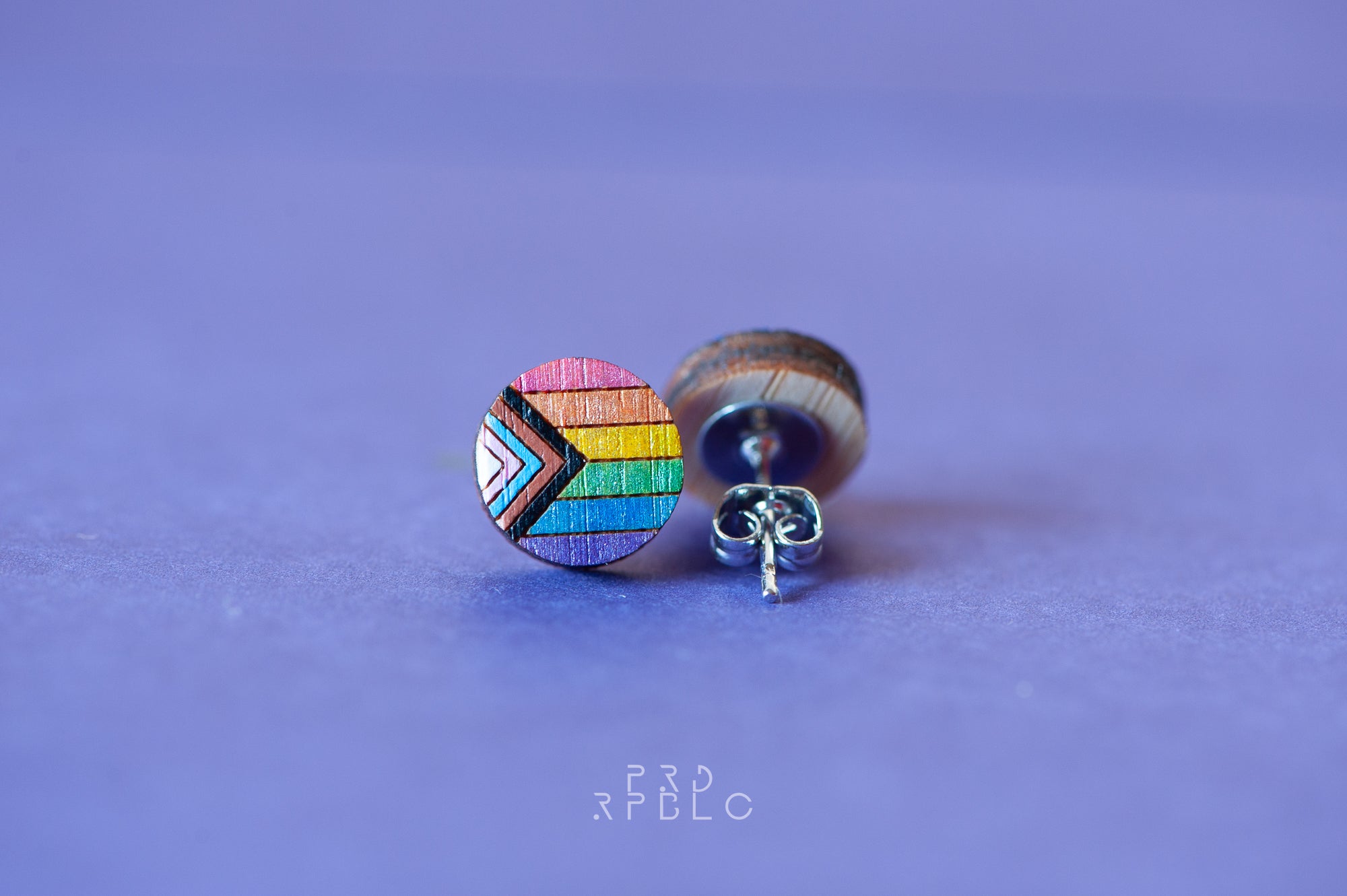A pair of round bamboo earrings featuring a glittered Progress Pride flag design, showcased against a blue backdrop, displaying both the front and back sides of the earrings.
