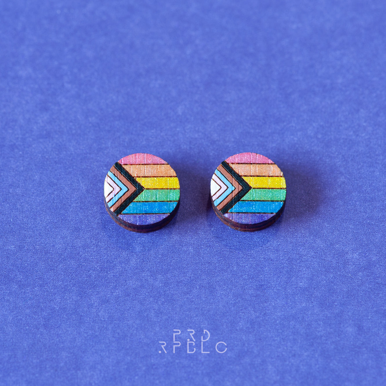 A pair of round bamboo earrings featuring a glittered Progress Pride flag design, showcased flat against the blue backdrop.