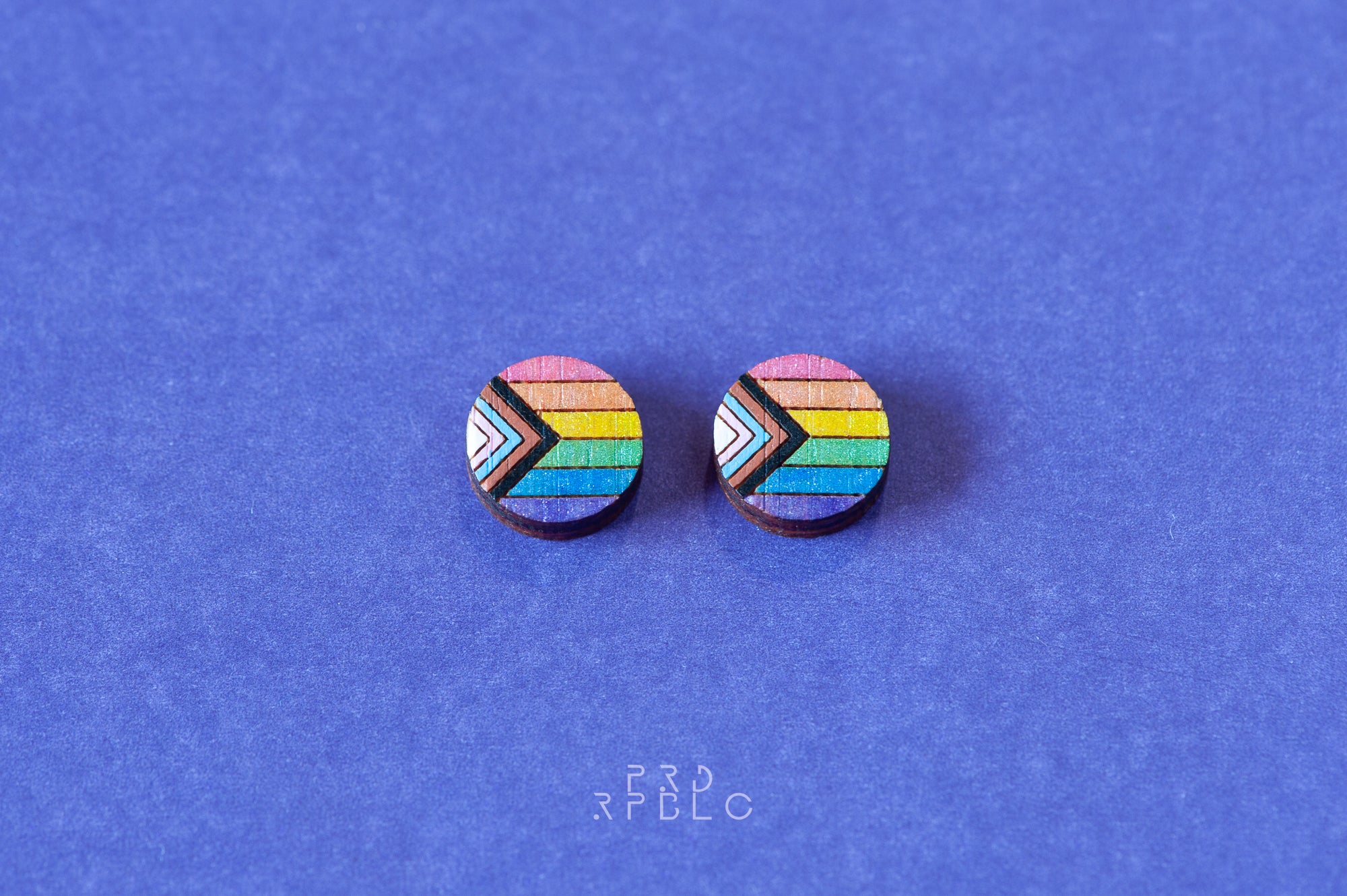 A pair of round bamboo earrings featuring a glittered Progress Pride flag design, showcased flat against the blue backdrop.