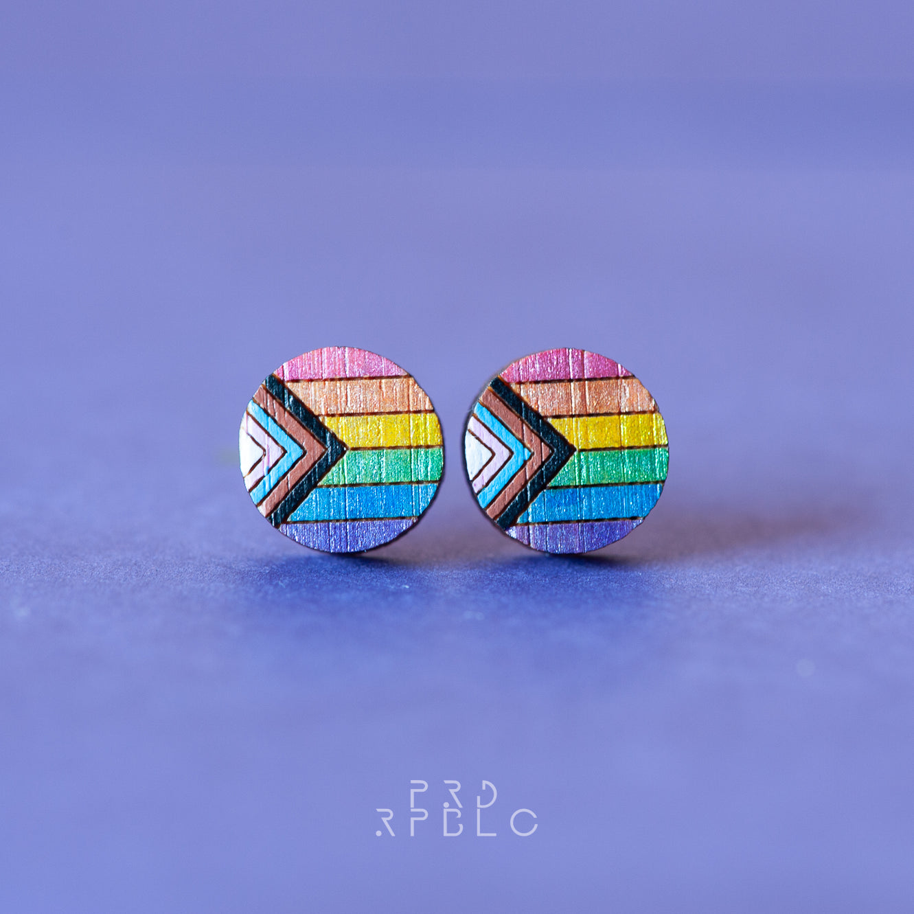 A pair of round bamboo earrings featuring a glittered Progress Pride flag design, showcased against a blue backdrop.