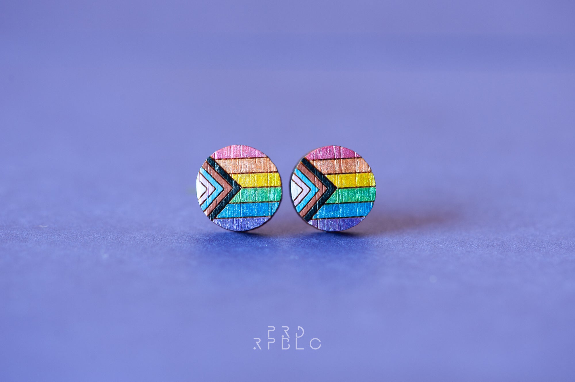 A pair of round bamboo earrings featuring a glittered Progress Pride flag design, showcased against a blue backdrop.