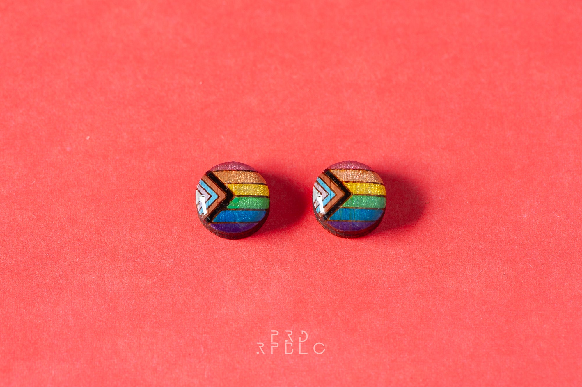 Round, glitter progress pride flag bamboo stud earrings on a red backdrop - earrings lying flat